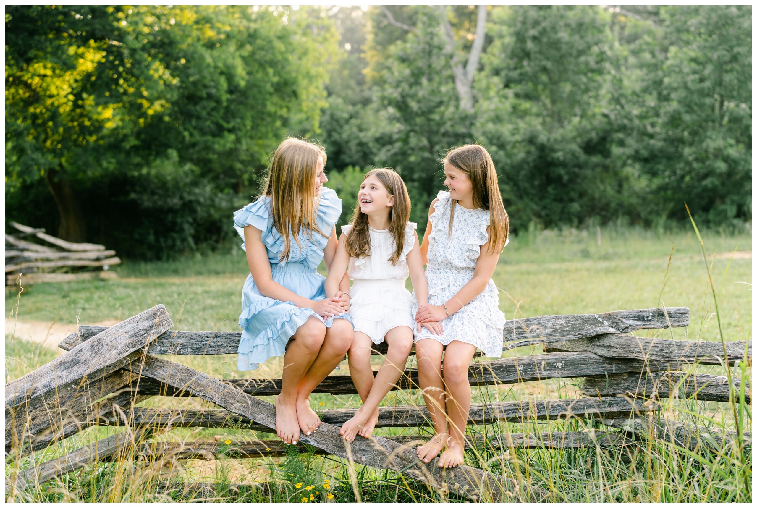 Portrait of three sisters sitting on a wooden fence smiling at one another.