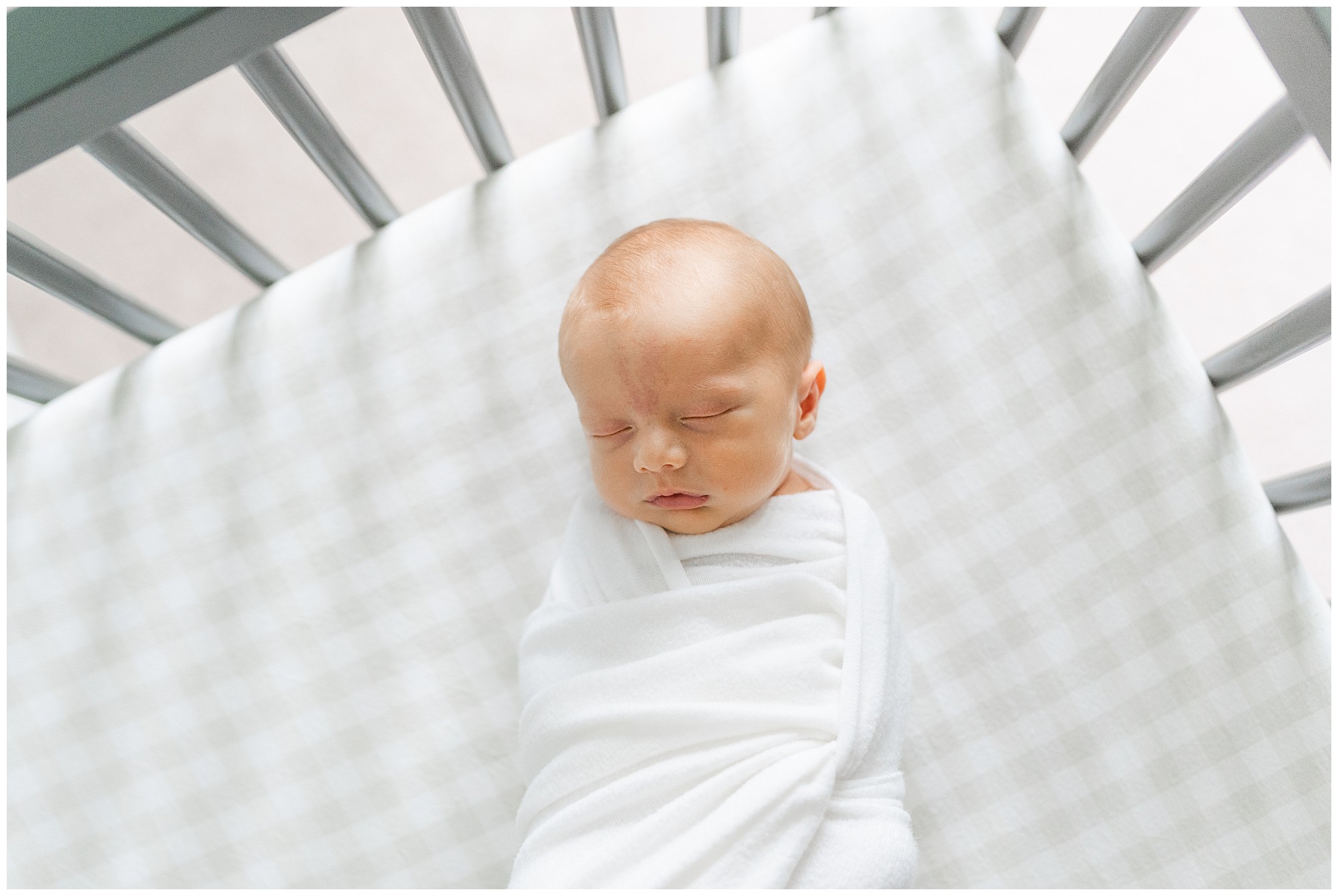 A sleeping baby wrapped in a white swaddle laying on a grey checkered crib sheet during an Atlanta in home newborn session.