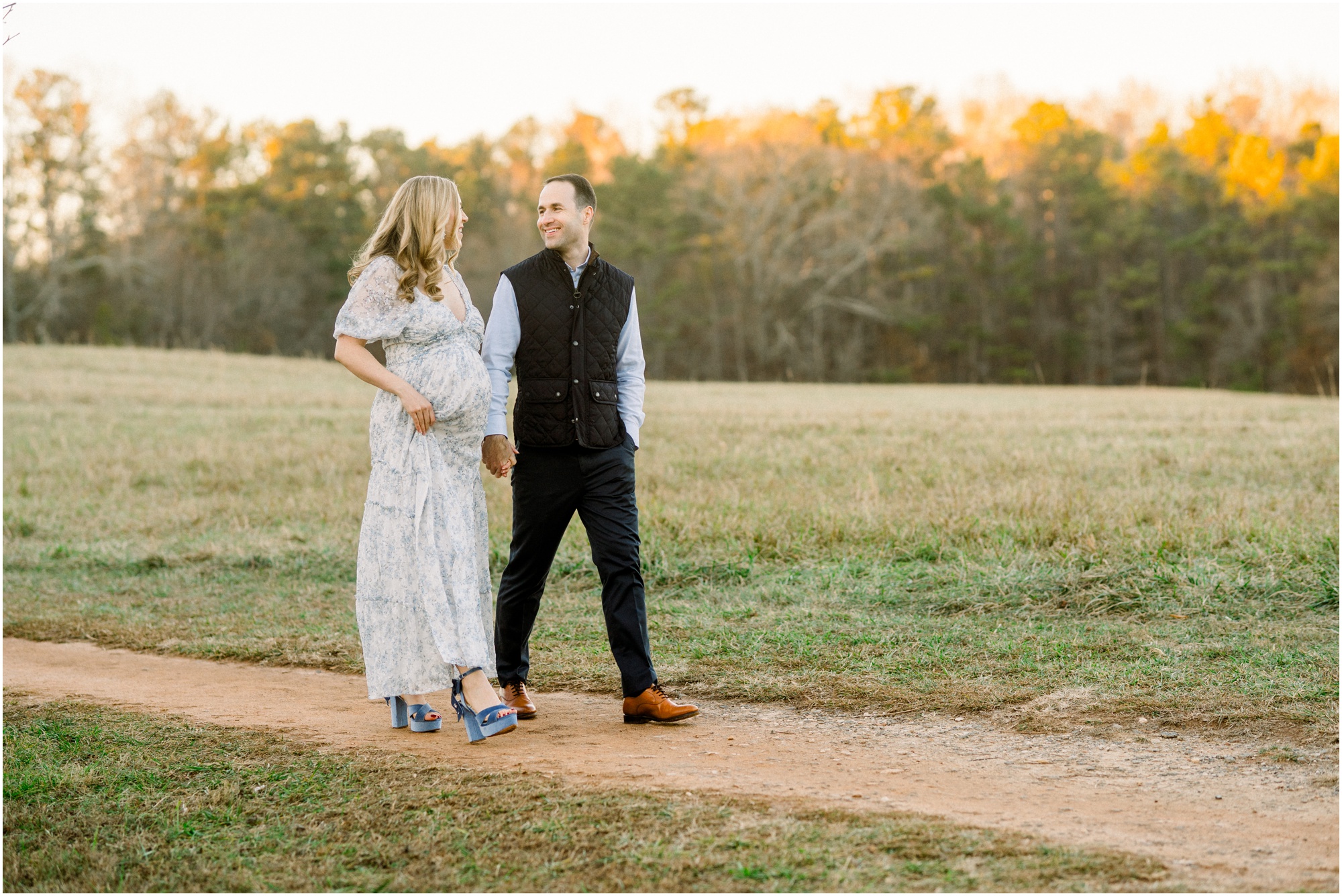 A pregnant woman holds her husbands hand and they smile towards each other as they walk down a dirt path in a green field for Atlanta maternity photos.